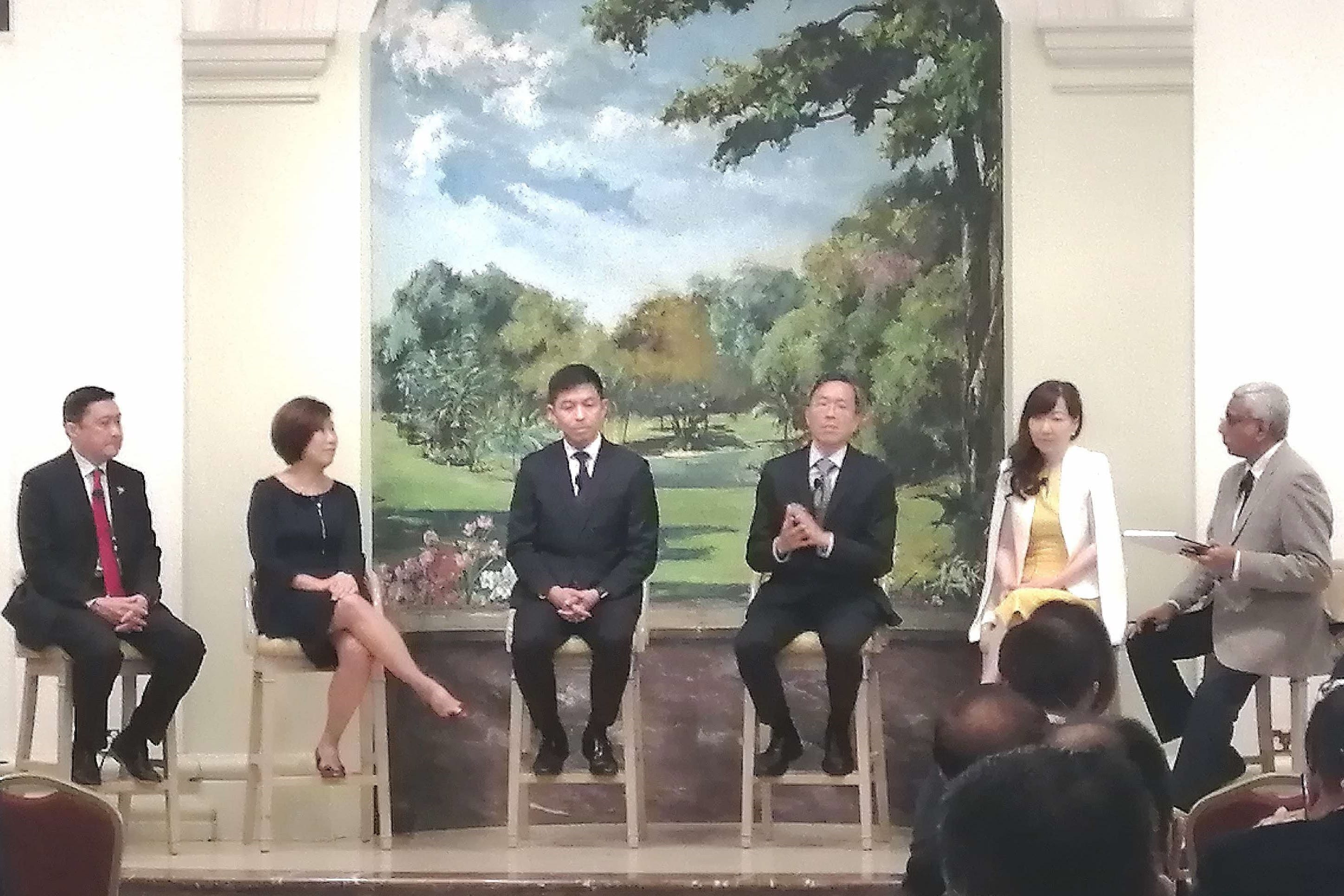 Six people seated on stools on a stage before a large landscape mural, facing an audience during a panel discussion.